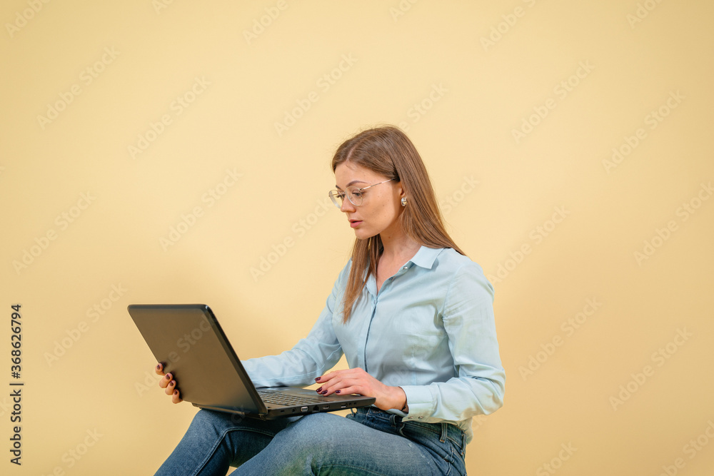 Young businesswoman using laptop over yellow background. Wearing in blue shirt and glasses. Looking at screen