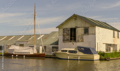 Old weathered boathouse with peeling paint on waterside, with moored wherry and pleasure boat. Blue sky with wispy cloud. Reflections in moving water. Landscape image, England.