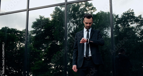 A man in a suit stands next to glass windows after rain and looks at his wristwatch.