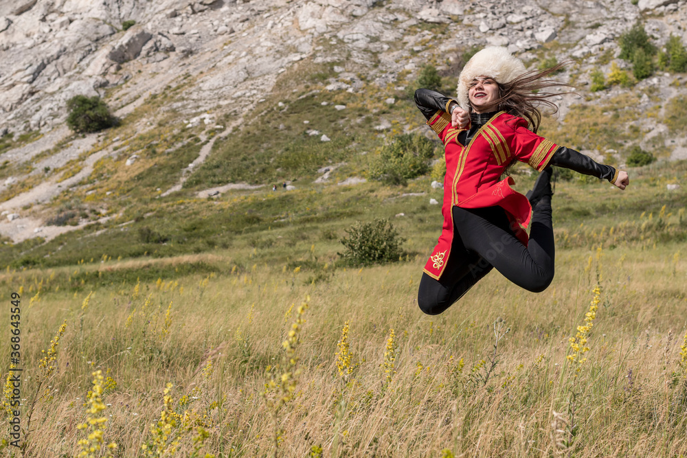 A cheerful young girl in a red suit soars over a green field, dancing a national dance on a mountain background