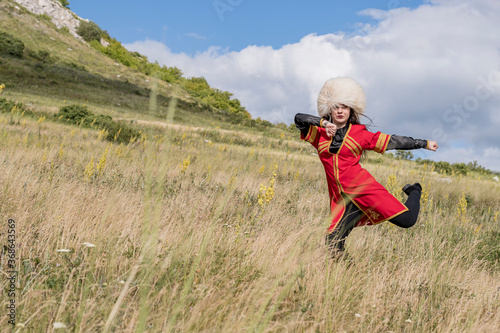 A young cheerful girl in a national costume is dancing a fiery dance on a green field at the foot of the mountain