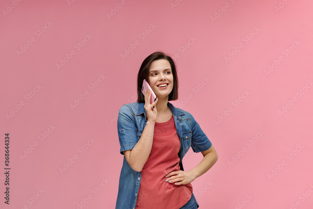 Young people working with mobile devices. Portrait of cute happy brunette woman wearing t-shirt and jeans shirt talking on mobile phone and smiling isolated over pink background. Copy space. Young people working with mobile devices. Portrait of cute happy brunette woman wearing t-shirt and jeans shirt talking on mobile phone and smiling isolated over pink background. Copy space.
