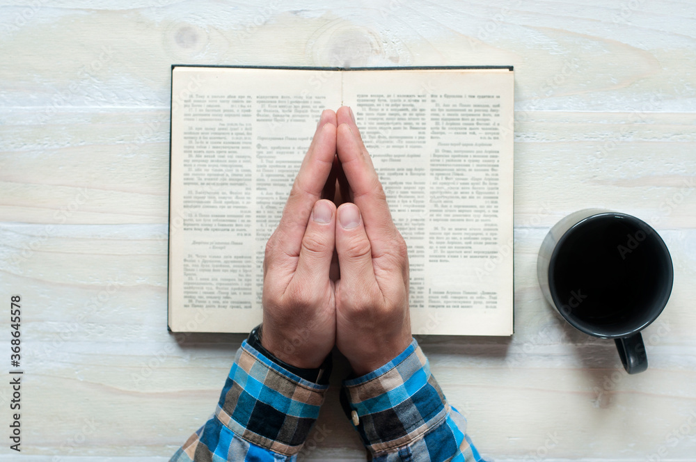 Naklejka premium Open Holy Bible. Hands folded in prayer. Cup on a wooden background.