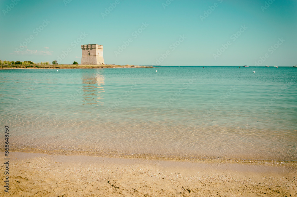 Spiaggia con mare trasparente, torre costiera e riflesso della torre sul mare.