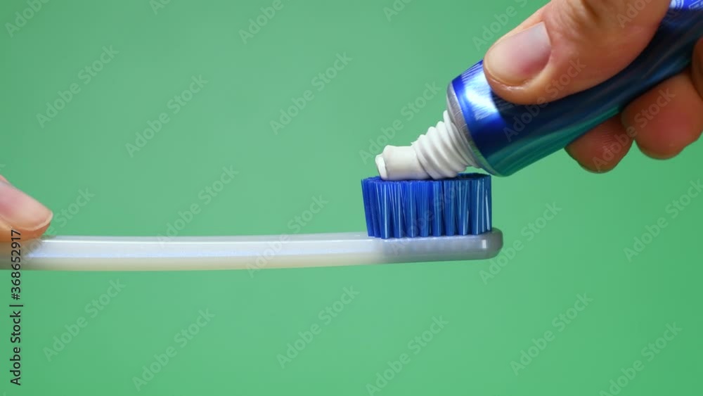 Female hand squeezing tooth paste on blue toothbrush. Green background.