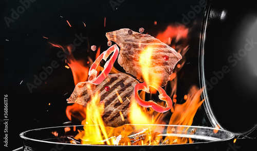 Meat and bell pepper slices falling onto barbecue grill with flame against black background, closeup