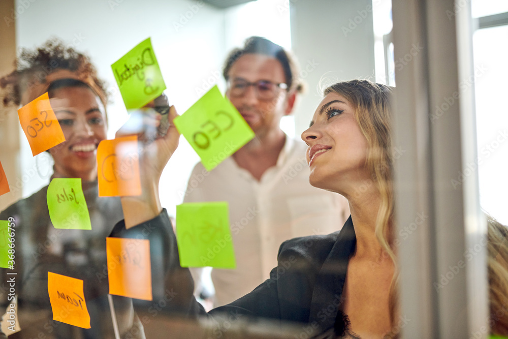 Smiling confident young businesswoman writing on adhesive note while discussing strategy with colleagues in board room during meeting Smiling confident young businesswoman writing on adhesive note while discussing strategy with colleagues in board room during meeting