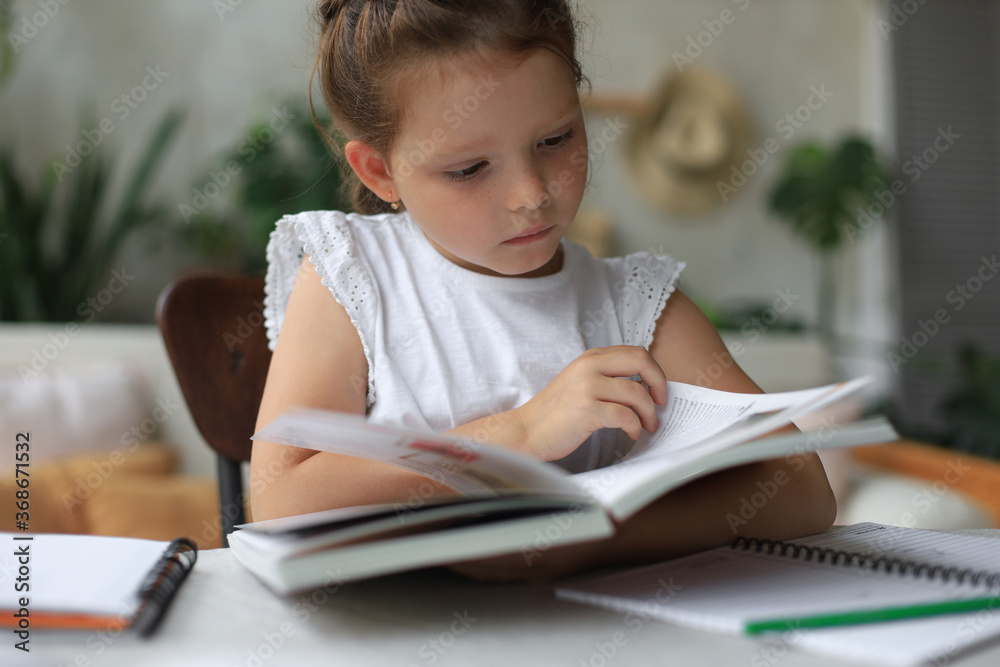 Little girl sits at the desl at home and does homework, self study of the material, reading book. The concept of distance education. Little girl sits at the desl at home and does homework, self study of the material, reading book. The concept of distance education.