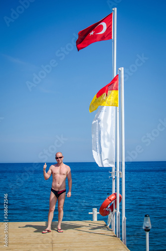 Handsome happy Caucasian man tourist stands on a pier on Mediterranean coast and smiles happily. Shows class. Happy summer and beach lifestyle in Turkey. Vacation Travel to sea. Last Minute Holidays