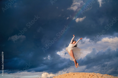 young ballerina in a light long white dress flies in a jump like a bird  a white skirt develops against the backdrop of a stormy evening sky. A rainbow appears among the clouds.