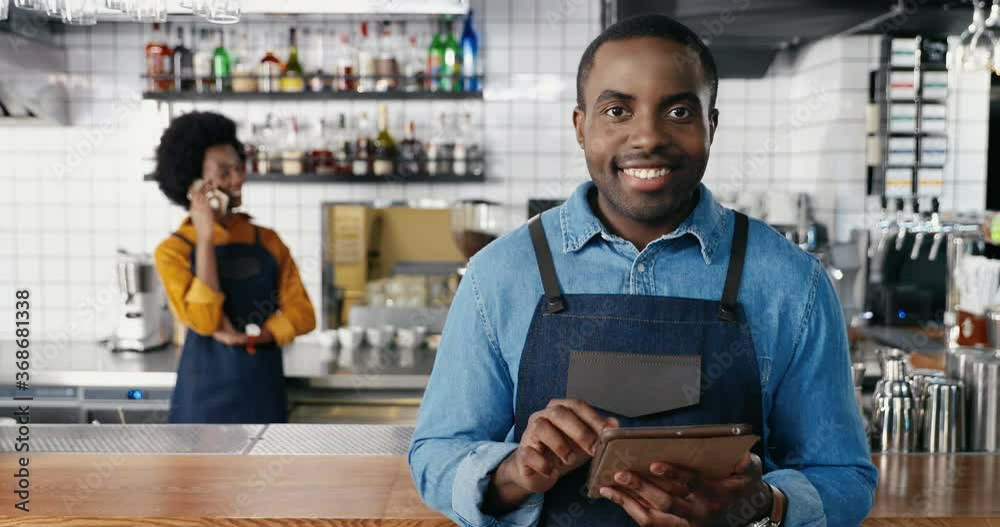 Portrait of male African American barmen using tablet device in cafe and smiling to camera. Handsome man waiter tapping and scrolling online on gadget. Waitress talking on mobile phone on background. Portrait of male African American barmen using tablet device in cafe and smiling to camera. Handsome man waiter tapping and scrolling online on gadget. Waitress talking on mobile phone on background.