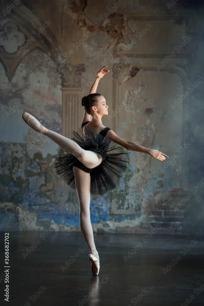 Portrait of a young ballerina on pointe shoes in a black swimsuit, tutu and pointe shoes in a luxurious interior. Stands in arabesque pose.