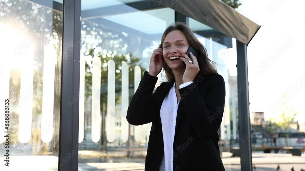 Attractive woman in white shirt and black jacket having conversation on smartphone while standing on street. Smiling female with dark hair talking on mobile with business partners. Attractive woman in white shirt and black jacket having conversation on smartphone while standing on street. Smiling female with dark hair talking on mobile with business partners.