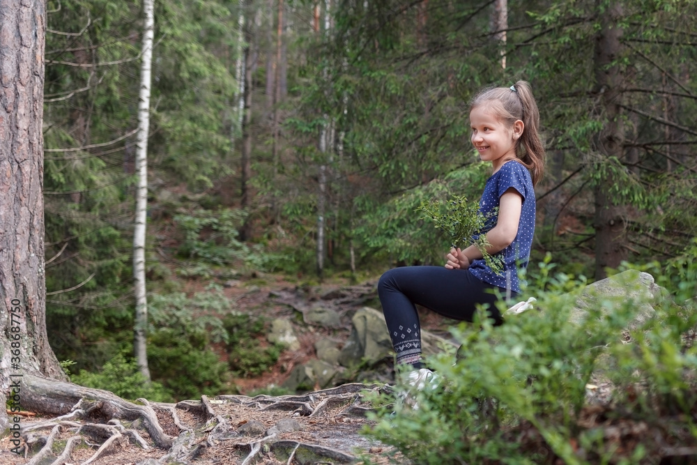 The girl in a purple T-shirt in the woods with a sprig of blueberries sat on a stone and smiles The girl in a purple T-shirt in the woods with a sprig of blueberries sat on a stone and smiles