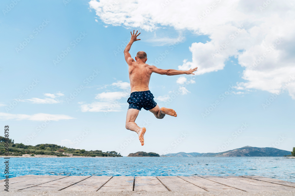 Φωτογραφία Middle-aged man dressed swimming trunks funny jumping to the waves from the boat pier as he having fun on merry vacation days on Adriatic sea coast