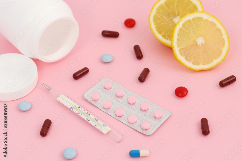 Multi-colored tablets in capsules on a light pink background. Thermometer, lemons and a white container for tablets.