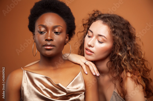two pretty girls african and caucasian blond posing cheerful together on browm background, etnithity diverse lifestyle people concept