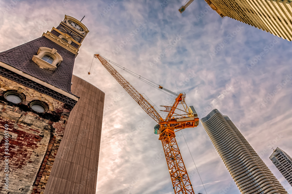 Looking up at a crane between a clock tower and a skyscraper.`