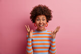 Excited cheerful woman with Afro hair keeps hands raised, hears awesome news, gets unexpected surprise, expresses joy and wonder, wears casual striped sweater, isolated on pink studio background