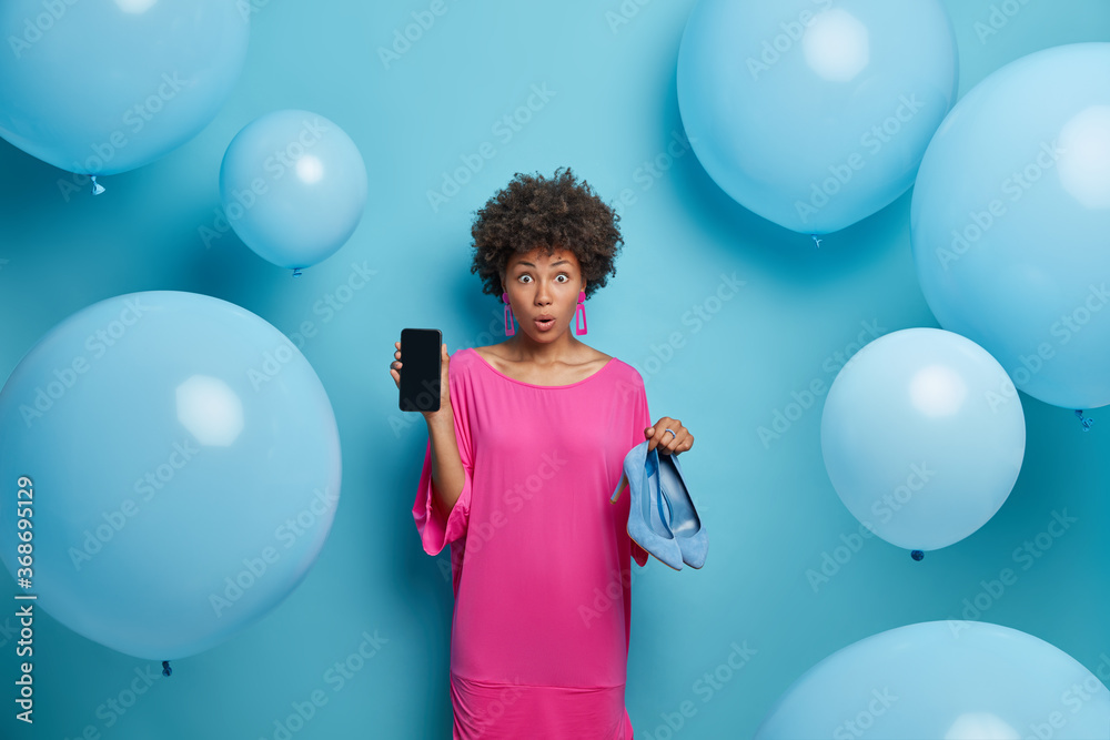 Fototapeta premium Photo of astonished curly young woman shows mobile phone display and high heeled shoes, makes shopping online, buys clothes in internet store, stands against blue background with balloons around