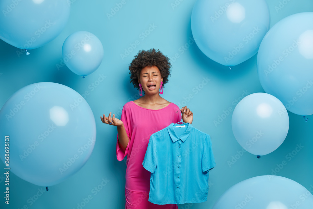 Fototapeta premium Womens fashion collection concept. Depressed crying dark skinned woman organizes clothes in wardrobe, holds blue shirt on hangers, has bad mood, thinks what to wear for party, blue background