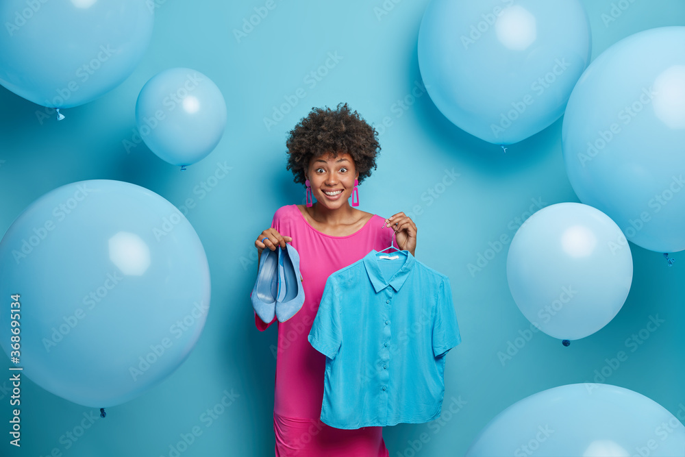Fototapeta premium Clothing, wardrobe and style concept. Happy ethnic woman chooses outfit to wear, holds high heeled shoes, dresses and prepares for date, expresses true positive emotions, stands indoor, decorated room