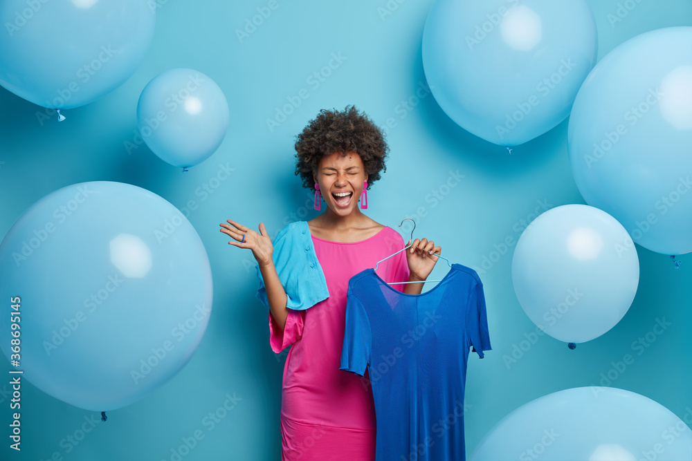 Emotional overjoyed dark skinned lady happy to have occasion for wearing favorite outfit, poses with long blue dress on hanger, has excellent mood, enjoys celebration and holiday preparation. Fashion Emotional overjoyed dark skinned lady happy to have occasion for wearing favorite outfit, poses with long blue dress on hanger, has excellent mood, enjoys celebration and holiday preparation. Fashion