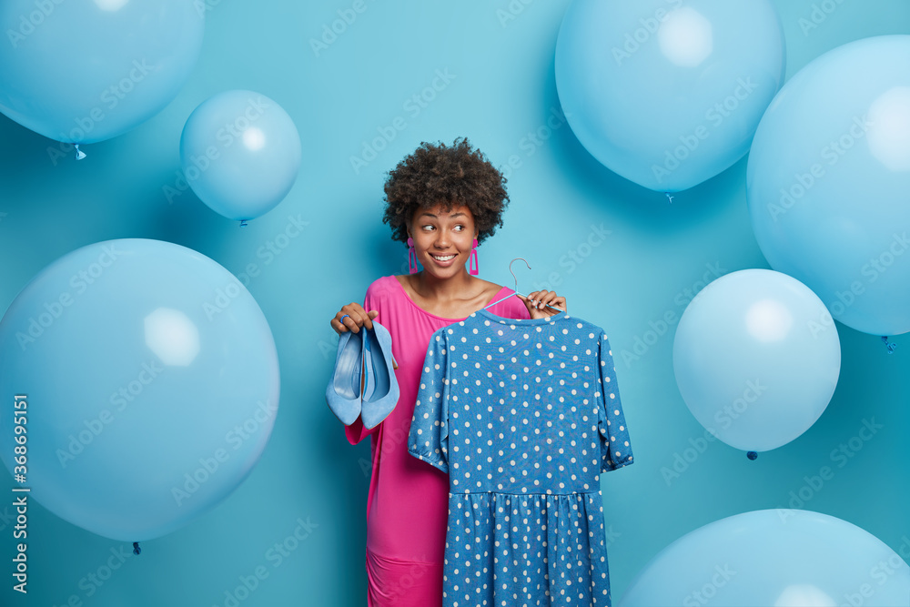 Fototapeta premium Style, clothes concept. Fashionable woman buys outfit for special occasion, waits party, holds fancy polka dot dress on hangers and blue high heeled shoes, surrounded by big inflated balloons