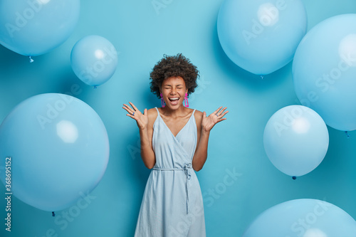 Shot of upbeat cheerful dark skinned woman feels very happy and excited, raises palms and laughs, spends free time on party, wears nice blue summer dress with earrings and rings, poses near balloons