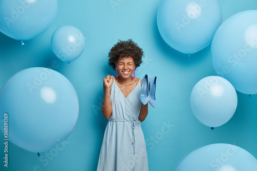 Women, fashion, clothes concept. Glad young African American woman clenches fist with joy, rejoices new purchase, buys fashionable outfit and shoes to dress for special occasion, blue color prevails