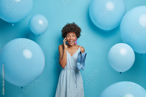 Beautiful winsome joyous woman organizes and prepares party event, invites friends via smart phone, chooses outfit to look brilliant, dressed in long dress and holds blue shoes, celebrates birthday