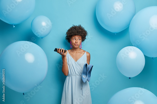 Lovely attractive thoughtful young Afro American woman prepares for date, holds smartphone in hand and waits for call from boyfriend, dresses blue dress and shoes, stands indoor near balloons