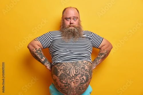 Confident serious blue eyed man with beard, has big abdomen, leads unhealthy lifestyle, dressed in striped undersized sailor t shirt, poses over yellow background. Plump guy stands self assured indoor