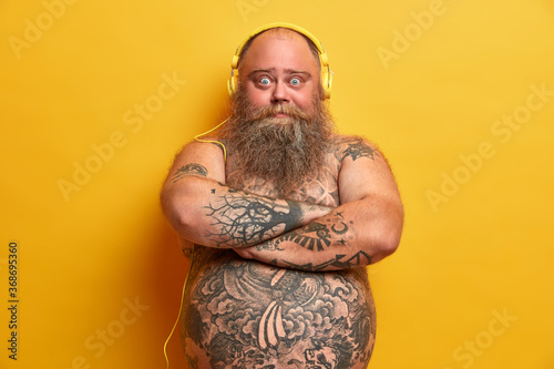 Chubby male meloman stands with arms folded, looks confident at camera, has tattooed body, listens music in headphones, thick beard and mustache, big fat belly, isolated on yellow background