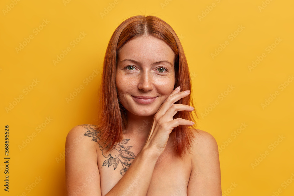 Natural beauty, purity and wellness concept. Satisfied redhead woman touches face gently, demonstrates her perfect smooth skin after spa procedure, stands bare shoulders against yellow studio wall Natural beauty, purity and wellness concept. Satisfied redhead woman touches face gently, demonstrates her perfect smooth skin after spa procedure, stands bare shoulders against yellow studio wall
