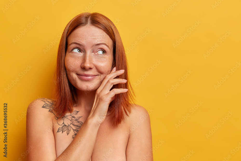 Fototapeta premium Ładna, rudowłosa młoda kobieta delikatnie dotyka policzka, patrzy z rozmarzonym zamyśleniem na bok, stoi naga, ma zadbane ciało i zdrową skórę, modelki na żółtym tle, pusta przestrzeń