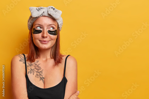 Horizontal shot of lovely adult woman dressed in sleevless black t shirt, has tattoo on body, looks away with thoughtful expression, wears headband and hydrogel patches, cares about skin and beauty photo