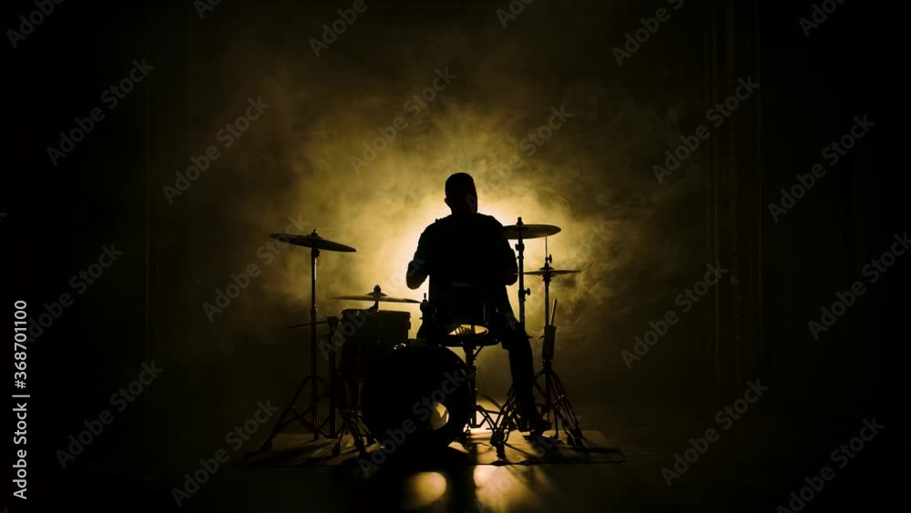 Silhouette drummer playing on drum kit on stage in a dark studio with smoke and neon lighting. Performance vocal and musical band. Slow motion.