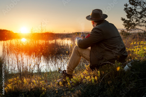 Male traveler with hat sitting in the bog and drinking hot drink from thermos during summer sunset