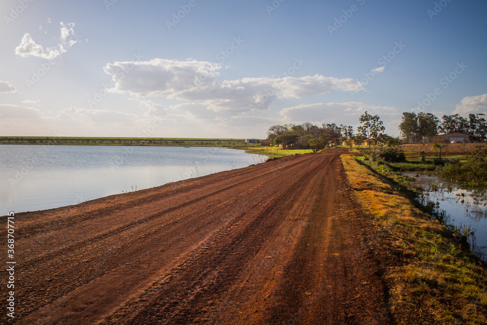 Naklejka premium Rural dirt road crossing a reservoir at sunset in the interior of Goiás, showcasing the beauty of the landscape with warm golden light reflecting off the water