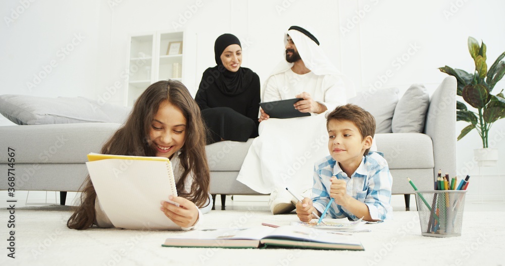 Arabian happy kids sitting on floor and painting in notebooks. Small sister and brother drawing at home. Couple of parents sitting on couch and talking. Arabs mother and father with tablet device. Arabian happy kids sitting on floor and painting in notebooks. Small sister and brother drawing at home. Couple of parents sitting on couch and talking. Arabs mother and father with tablet device.