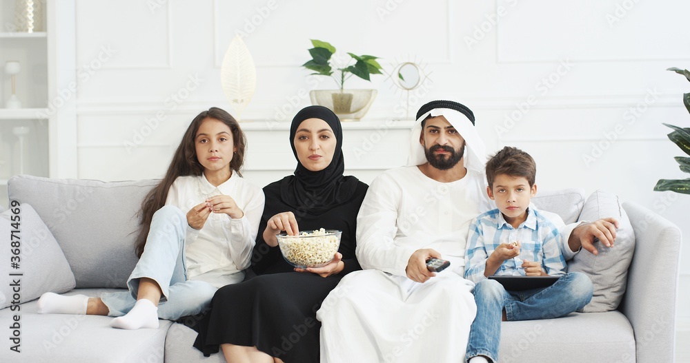 Cheerful Arabian parents with little children sitting on couch, smiling, watching movie on TV and eating popcorn. Cute Arab small kids with mother and father spending time together at home.