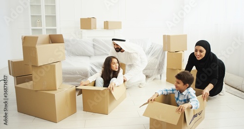 Cheerful young muslims parents in hijab and dishdasha playing with kids in lving room while moving in new home. Arabs mother and father riding small son and daughter in carton boxes.