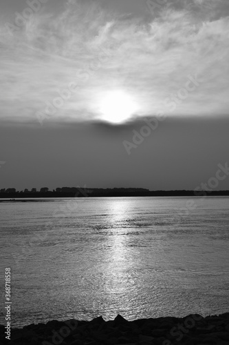 Black and white sunset. Reflection of sunlight in the river. The sun in a cloud cover. Outlines of rocks on the shore. Vertical photo.