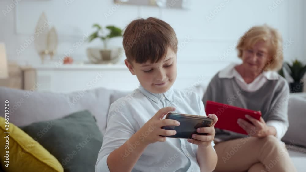 Joyful young teenager boy kid playing games on online smartphone. His grandmother relaxing on sofa watching movies online on tablet. Family staying home. Joyful young teenager boy kid playing games on online smartphone. His grandmother relaxing on sofa watching movies online on tablet. Family staying home.
