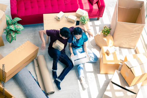 Overhead view of young Asian couple relax together after moving house. Young man and woman sitting on the floor, using laptop against red sofa with many boxes in living room.