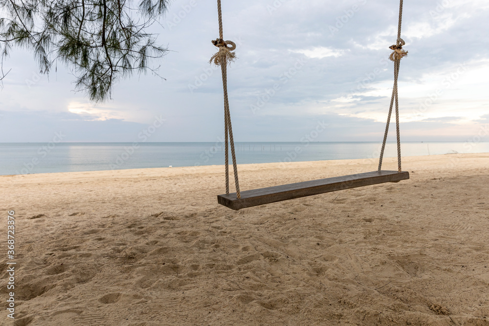 Little wooden swing under shade of big trees at peaceful beach on late afternoon. Blue cloudy sky and calm sea Little wooden swing under shade of big trees at peaceful beach on late afternoon. Blue cloudy sky and calm sea