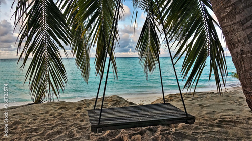 Holidays in the Maldives. The swing is suspended from a palm tree on the beach. Through the leaves you can see the aquamarine ocean  the sky with picturesque clouds. There are footprints in the sand. 