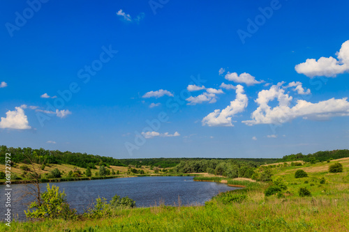Summer landscape with beautiful lake, green meadows, hills, trees and blue sky © olyasolodenko