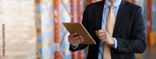 Close up businessman in black suit investor using tablet checking inventory in warehouse industry, panorama banner size.